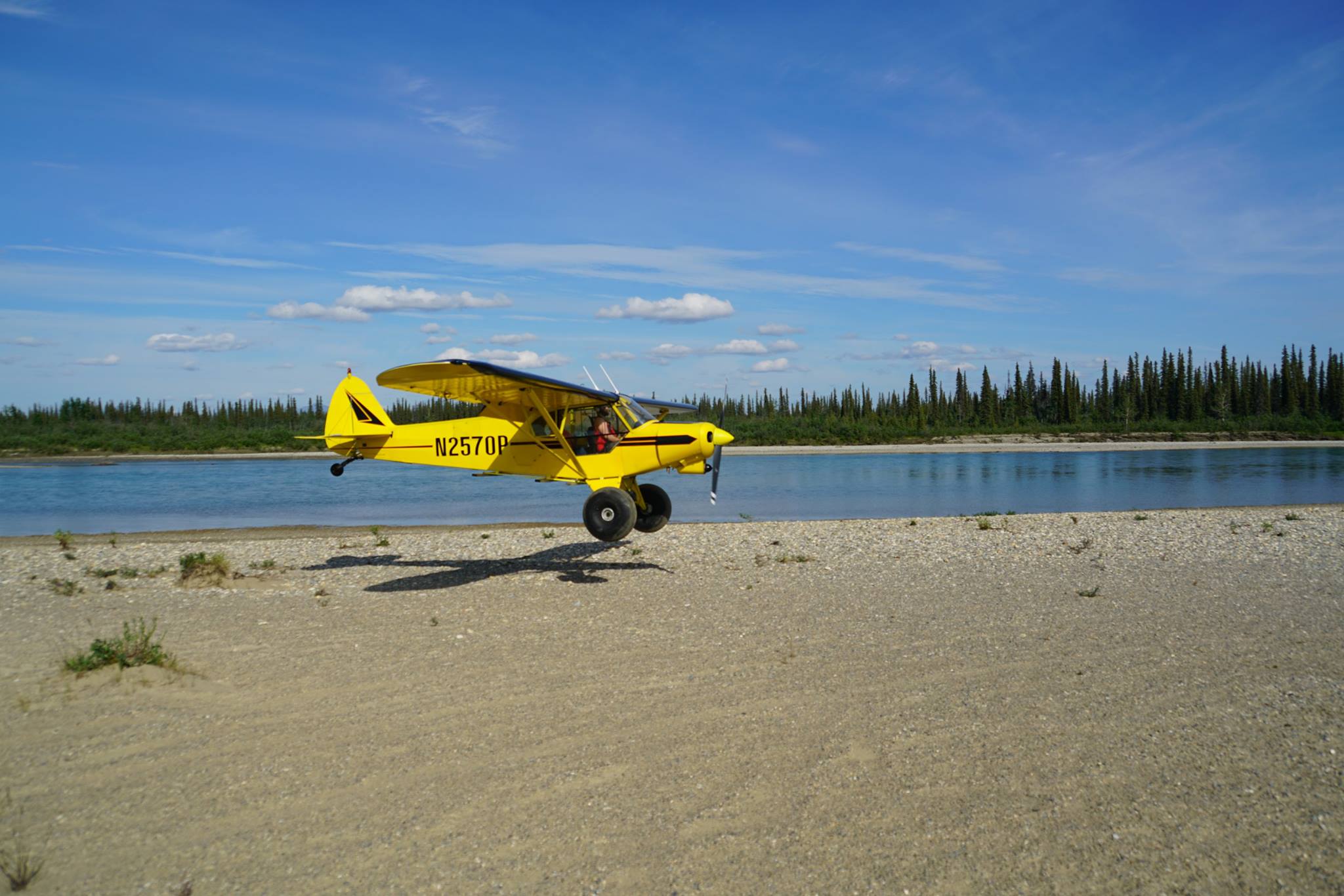 A yellow bush plane flying low above a gravel riverbank in Alaska